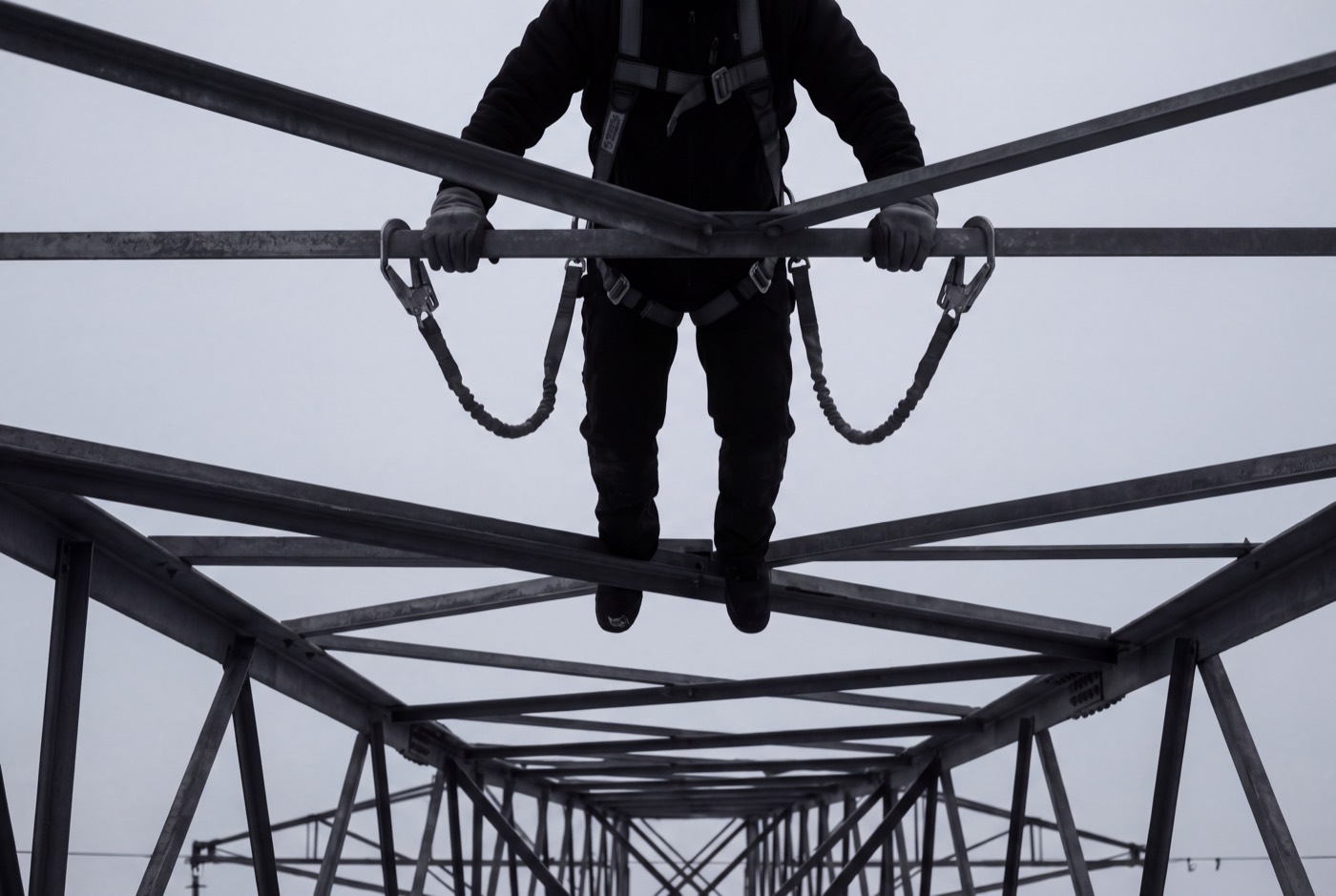 A harnessed worker with twin shock-absorbing lanyards standing on the steel lattice of a transmission tower against a pale sky
